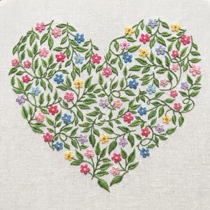 May include: Embroidered heart design with colourful flowers in pink, blue, yellow, and purple, surrounded by green leaves. The floral pattern is stitched on a white fabric background, creating a handcrafted appearance.