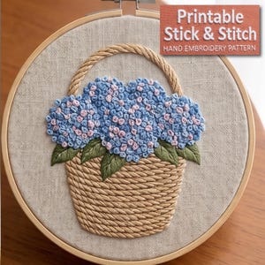 May include: A round wooden embroidery hoop containing a beige fabric panel with a hand-stitched design of a tan rope basket filled with blue and light pink hydrangea flowers and green leaves. Text in the corner reads Printable Stick & Stitch.