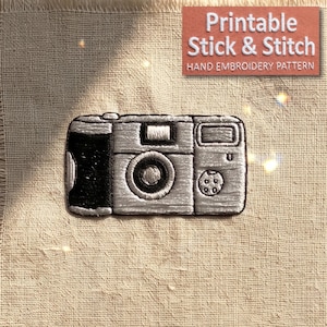 May include: An embroidered camera design on beige fabric featuring a black grip, a central lens, and a flash unit. Above the camera, a rectangular label displays the white text: Printable Stick & Stitch HAND EMBROIDERY PATTERN.