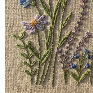 Peut inclure: Gros plan d'une broderie florale. Le motif pr&eacute;sente des fleurs bleues, violettes et jaunes avec des tiges et des feuilles vertes. La broderie est r&eacute;alis&eacute;e sur un fond de tissu beige, mettant en valeur la texture du mat&eacute;riau.
