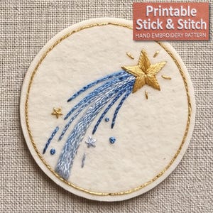 May include: A circular off-white fabric patch featuring an embroidered gold shooting star with a trailing tail of blue and silver thread. The design includes small embroidered stars and dots, all enclosed within a thin gold circular border.