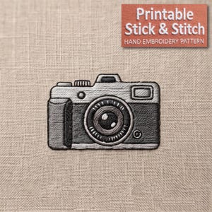 May include: An embroidered patch of a camera in shades of gray and black on a light beige fabric background. The design features a central lens, a shutter button, and a viewfinder. The text 'Printable Stick & Stitch HAND EMBROIDERY PATTERN' is visible.