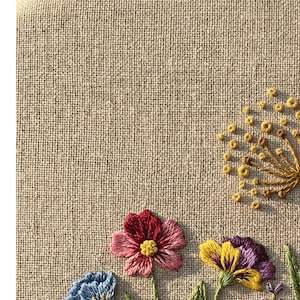 Peut inclure: Gros plan sur un tissu de lin beige avec une broderie florale color&eacute;e. La broderie pr&eacute;sente une fleur rouge et rose, une fleur jaune et violette, et une fleur bleue. Le motif comprend &eacute;galement des feuilles vertes et un pissenlit jaune.