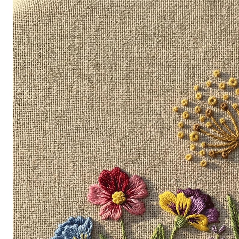 Peut inclure: Gros plan sur un tissu de lin beige avec une broderie florale color&eacute;e. La broderie pr&eacute;sente une fleur rouge et rose, une fleur jaune et violette, et une fleur bleue. Le motif comprend &eacute;galement des feuilles vertes et un pissenlit jaune.