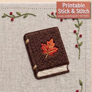 Könnte beinhalten: Ein brauner, gestickter Buch-Aufnäher mit einem zentralen orange-roten Ahornblatt-Motiv, cremefarbenen Seitenkanten und Buchrücken-Details. Der Text "Printable Stick & Stitch HAND EMBROIDERY PATTERN" ist in der oberen rechten Ecke zu sehen.