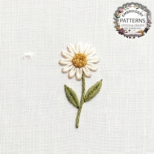 Peut inclure: Une marguerite brodée à la main sur un tissu blanc avec des pétales blancs, un cœur jaune texturé, deux feuilles vertes et une tige verte. Un logo circulaire en haut à droite indique EMBROIDERY PATTERNS STITCH & CREATE PDF DOWNLOAD.