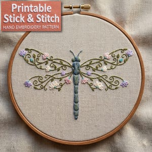 Puede incluir: Diseño de libélula bordada a mano sobre tela de lino, en un bastidor de madera. La libélula presenta detalles florales en verde, rosa y morado. El texto "Printable Stick & Stitch" es visible, indicando un patrón de bordado.