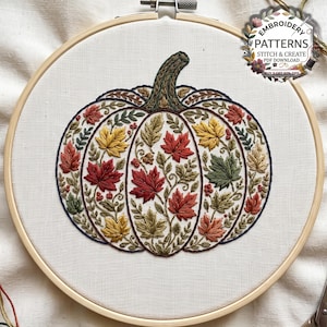 May include: An embroidery hoop containing a white fabric panel with a stitched pumpkin design. The pumpkin is filled with detailed red, yellow, and green maple leaves and small berries. Text in the corner reads EMBROIDERY PATTERNS.