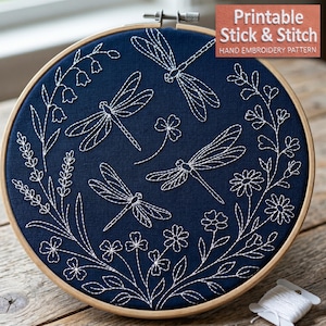 Könnte beinhalten: Ein runder Stickrahmen aus Holz mit dunkelblauem Stoff, auf dem drei Libellen sowie verschiedene Blumen und Blätter in weißen Linien gestickt sind. Der Text "Printable Stick & Stitch HAND EMBROIDERY PATTERN" ist oben rechts zu sehen.