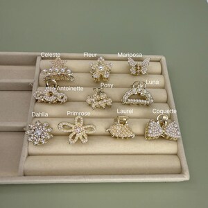 May include: A selection of gold-coloured hair claw clips in diverse designs, including floral, butterfly, and bow shapes. Each clip is adorned with pearls and crystals. Names such as "Celeste," "Fleur," and "Mariposa" are visible.