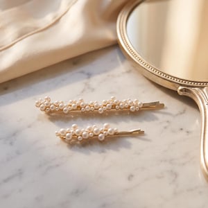 May include: Two gold-toned hair clips decorated with clusters of small white faux pearls of varying sizes. The hair accessories are arranged horizontally, featuring a slender metal base and a secure slide-in design for styling hair.