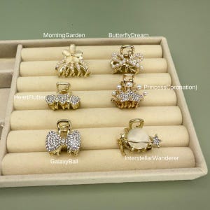 Könnte beinhalten: Eine Sammlung goldfarbener Haarspangen mit verschiedenen Designs, darunter Blumen-, Perlen- und Kristallverzierungen. Die Spangen sind in einer beigen Präsentationsschale angeordnet, mit Textetiketten wie "MorningGarden" und "GalaxyBall".