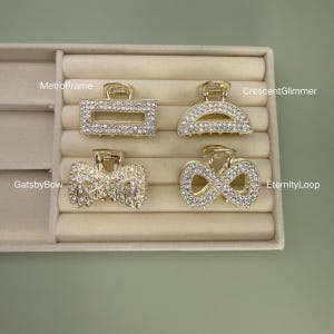 May include: Four gold-toned hair claw clips with rhinestone embellishments. The clips are in various shapes: a rectangle, a half-circle, a bow, and an infinity symbol. The clips are displayed in a beige jewelry tray.