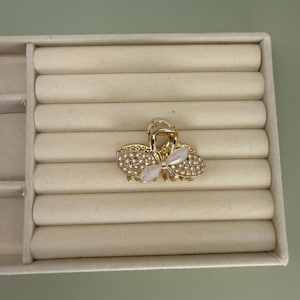 May include: A gold-toned hair claw clip with a butterfly design. The clip is encrusted with small, clear rhinestones and features a central butterfly-shaped accent with two white, oval-shaped stones.