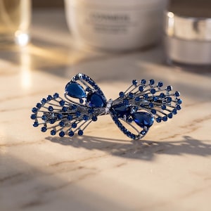 May include: A hair clip featuring a dark metal frame shaped like a bow, adorned with numerous small round blue rhinestones and four larger teardrop-shaped blue crystals. The intricate design includes thin metal wire branches extending outward.