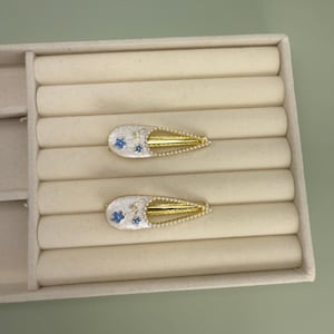 May include: Two decorative hair clips. Each clip features a white base with blue floral accents and a gold-colored metal frame. The clips are arranged on a beige, segmented surface, suggesting a jewelry display or storage.