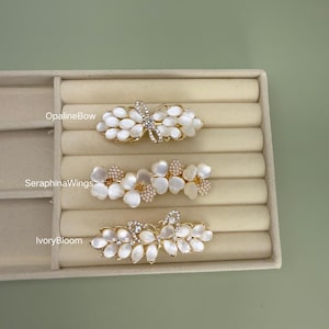 May include: Three decorative hair barrettes with floral and butterfly designs. The barrettes feature white, petal-shaped accents and gold-colored metal frames. One barrette has a butterfly design with clear crystal accents. The other two have floral designs with pearl and crystal accents.