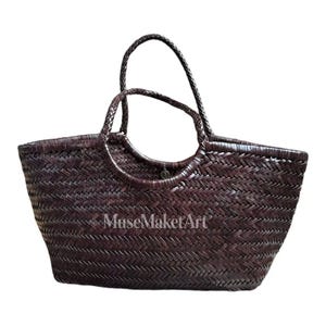 Puede incluir: Un bolso tote de cuero trenzado en color marrón oscuro con un patrón de espiga, dos asas circulares trenzadas y un pequeño dije metálico cerca del centro superior. El texto MuseMaketArt está impreso en blanco en el frente del bolso.