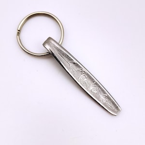 May include: A silver-toned metal keyring featuring a flat, elongated handle with a textured surface and an embossed floral pattern of three roses. The keyring includes a circular split ring attached to the top of the decorative metal piece.