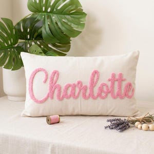 May include: A rectangular off-white throw pillow featuring the name Charlotte embroidered in a raised, textured pink script font. The pillow is positioned on a light-colored surface with a small spool of pink thread and dried lavender nearby.