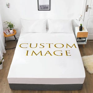 Può includere: Un lenzuolo con angoli bianco con il testo CUSTOM IMAGE stampato in grandi lettere dorate al centro. Il lenzuolo è disposto su un letto con due cuscini bianchi, in una stanza con pavimento in legno e un comodino.