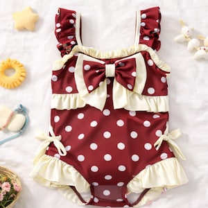 Può includere: Un costume da bagno bordeaux a pois con un grande fiocco color crema e dettagli a balze. Il costume intero ha balze color crema intorno alle gambe e un fiocco color crema sul davanti. Le spalline sono bordeaux.