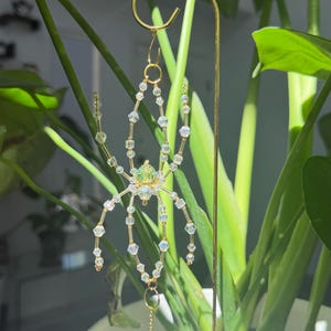 May include: A decorative spider ornament crafted from clear and light green faceted beads on a gold-toned wire frame. The spider hangs from a gold metal hook and features a dangling clear crystal prism attached to a circular gold ring at the bottom.