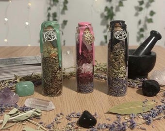 May include: Three glass bottles filled with herbs and crystals, sealed with colored wax and pentacle charms. The bottles are green, pink, and black. Various crystals, herbs, and a mortar and pestle surround the bottles on a wooden surface.