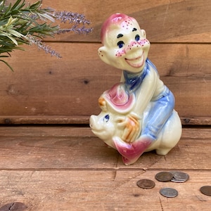 May include: A vintage ceramic figurine depicts a smiling figure riding a pig. The figure wears a pink hat and blue overalls. The pig is white with pink accents. The figurine is set against a wooden background.