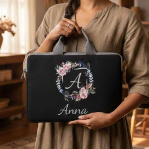 May include: Black laptop sleeve with a floral wreath design, the letter "A", and the name "Anna" in white script. The sleeve has a gray handle and zipper. The background is blurred, with a person holding the sleeve.