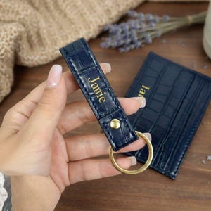 Può includere: Un portachiavi in pelle blu navy con finiture dorate e il nome "Jamie" impresso in lettere dorate. Un portacarte blu navy abbinato è accanto al portachiavi. Gli articoli hanno una trama a pelle di coccodrillo.