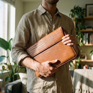 May include: A brown leather laptop case with a crocodile skin texture. The case is rectangular with a flap closure and a zipper. The case is held by a person wearing a tan button-down shirt.