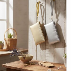 Puede incluir: Dos pequeñas bolsas de muñeca rectangulares, una dorada y otra plateada, cuelgan de ganchos de latón en una pared de madera blanca. Cada bolsa tiene una correa de muñeca a juego y un cierre de cremallera. Las bolsas se exhiben en un entorno rústico y bien iluminado.