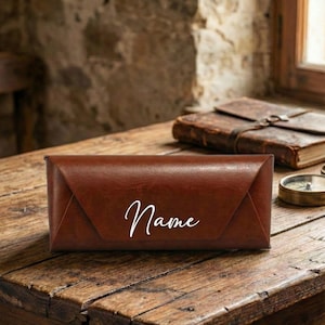 Puede incluir: Un estuche de gafas de cuero marrón con la palabra "Name" en escritura blanca. El estuche es rectangular con un cierre de solapa plegada. Está sobre una superficie de madera.
