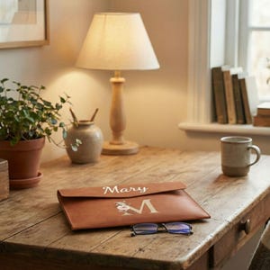 Puede incluir: Un portadocumentos de cuero marrón con el nombre "Mary" y un diseño floral "M" está sobre un escritorio de madera. Una lámpara de madera con pantalla beige, una planta en maceta, un jarrón de cerámica, una taza y gafas también están en el escritorio.