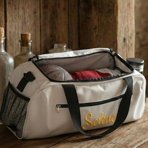 Könnte beinhalten: Eine hellgraue Sporttasche mit schwarzen Akzenten, einer Netztasche und einem Reißverschluss. Die Tasche hat schwarze Griffe und den Namen "Selene" in gelber Schrift gestickt. Im Inneren befinden sich ein Basketball und rote Kleidung.