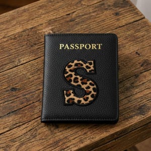 Könnte beinhalten: Schwarzer Reisepasshalter mit dem Wort „PASSPORT“ in goldfarbenen Buchstaben. Der Buchstabe „S“ ist in einem Leopardenmuster gehalten. Der Reisepasshalter liegt auf einer Holzoberfläche.