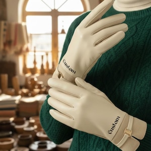 May include: Cream-colored gloves with the word "Custom" printed on the wrist cuffs. The gloves have a smooth leather appearance and a contrasting beige fabric detail at the wrist, secured with a gold-colored buckle. The person is wearing a green sweater.