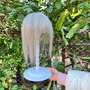 Könnte beinhalten: Eine silber-graue Perücke mit langem, glattem Haar wird auf einem weißen Perückenständer präsentiert. Der Ständer hat einen runden Sockel und einen schlanken, gebogenen Stiel. Der Hintergrund zeigt grünes Laub und einen Metallzaun.