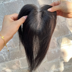 Puede incluir: Una pieza de cabello castaño oscuro con una raya de aspecto natural. La pieza de cabello está sujeta por dos manos, mostrando los finos mechones y la apariencia realista del cabello. La pieza de cabello está diseñada para agregar volumen y cobertura.