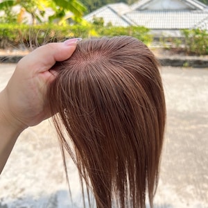 Op de afbeelding: Een bruine haartop met lange, rechte lokken. Het haarstuk wordt omhoog gehouden en toont de fijne mesh-basis en de natuurlijk ogende haarvezels. De kleur is een warm, medium bruin, geschikt om te mengen met verschillende haartinten.