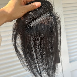 May include: A dark brown hairpiece with a fine mesh base. The hair is long and straight, with a natural shine. The hairpiece is held up by a hand, showing the attachment points.