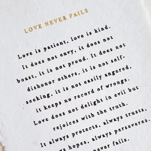 May include: A rectangular piece of white textured paper with deckled edges featuring black printed text that reads Love is patient, love is kind. It does not envy, it does not boast, it is not proud. It does not dishonor others, it is not self-seeking, it is not easily angered, it keeps no record of wrongs. Love does not delight in evil but rejoices with the truth. It always protects, always trusts, always hopes, always perseveres. Love never fails. The text is framed by the gold printed phrases LOVE NEVER FAILS at the top and 1 CORINTHIANS 13:4-8 at the bottom.