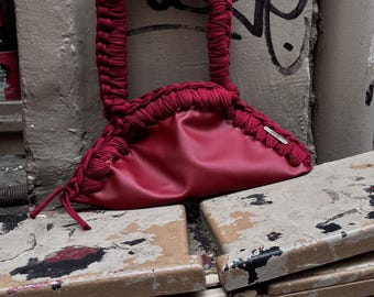 Puede incluir: Un bolso de mano rojo con asa trenzada y cuerpo fruncido. El bolso está hecho de un material liso y presenta una pequeña etiqueta plateada. El bolso está sobre una superficie desgastada con graffiti.