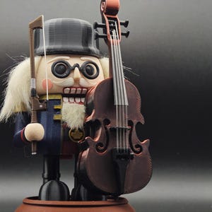 Peut inclure: Figurine décorative de casse-noisette avec un chapeau haut de forme noir, des lunettes et une barbe blanche. Le casse-noisette tient un violon et un archet miniatures. La figurine est peinte en bleu, rouge et or.