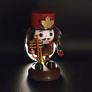 Peut inclure: Figurine en bois de casse-noisette, portant un uniforme et un chapeau rouges, tenant une trompette dorée. La figurine a un visage blanc avec une moustache et une bouche rouge. La base du casse-noisette indique "Ugly Things 3".