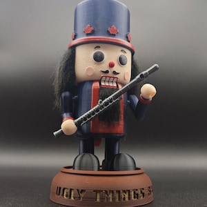 Peut inclure: Figurine décorative de casse-noisette avec un chapeau et un uniforme bleus, tenant une flûte noire. La figurine a un visage peint avec une moustache et des cheveux noirs. La base indique "UGLY THINGS".