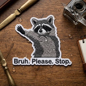 May include: Embroidered patch featuring a raccoon with a black and white face and gray fur, raising a paw. The patch has the text "Bruh. Please. Stop." in white letters on a white background. The patch is on a wooden surface.