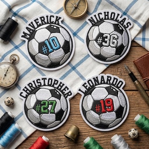 May include: Four soccer ball patches with personalized names and numbers. Each patch features a soccer ball design with a name above and a number below. The numbers are in different colors: blue, white, green, and red.