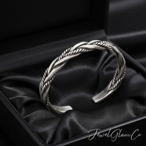 May include: A silver cuff bracelet with a braided design, presented in a black box with black satin lining. The bracelet features a textured, rope-like pattern. The overall aesthetic is elegant.